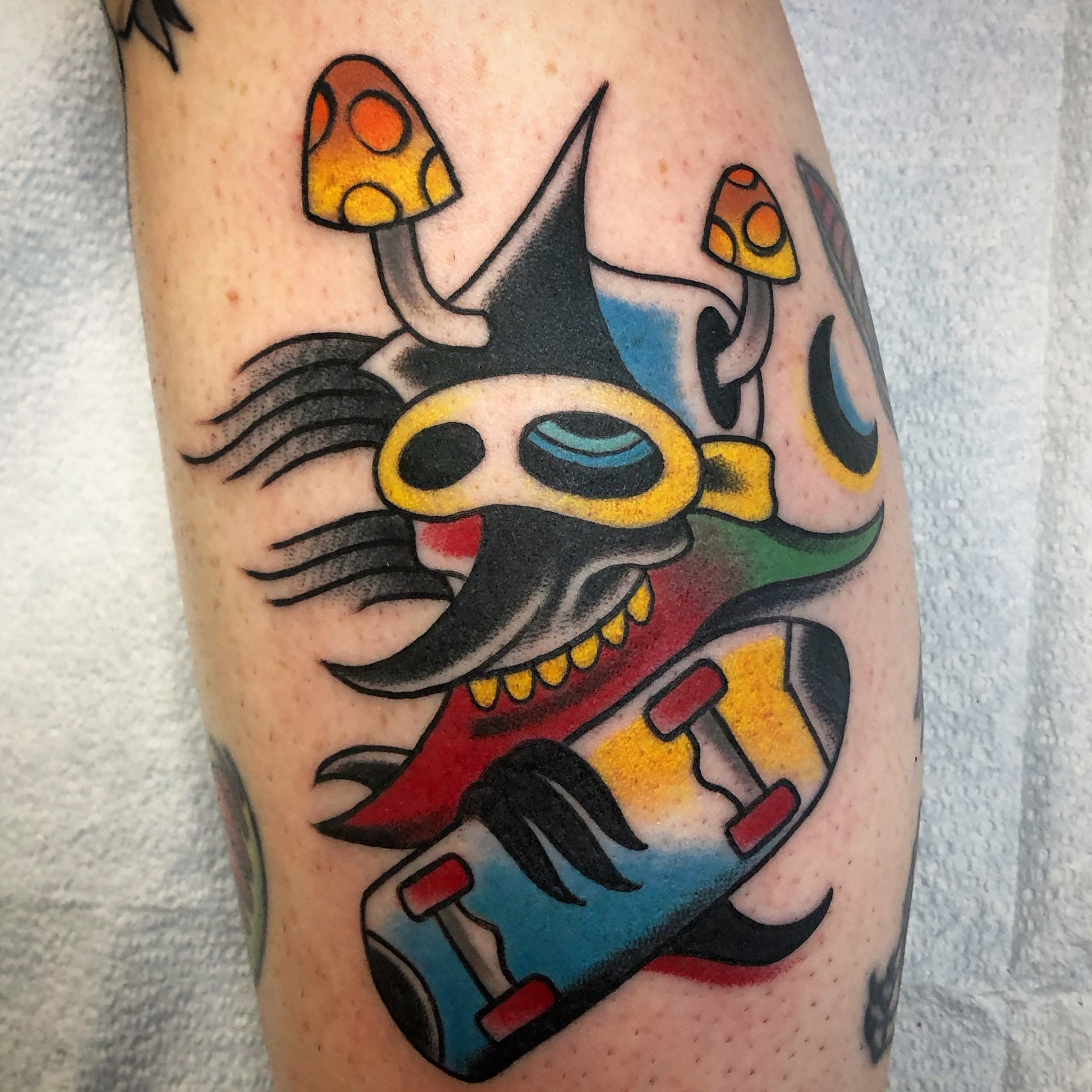 Colorful cartoon-style tattoo of a black creature with yellow goggles, mushrooms, and a skateboard on skin.