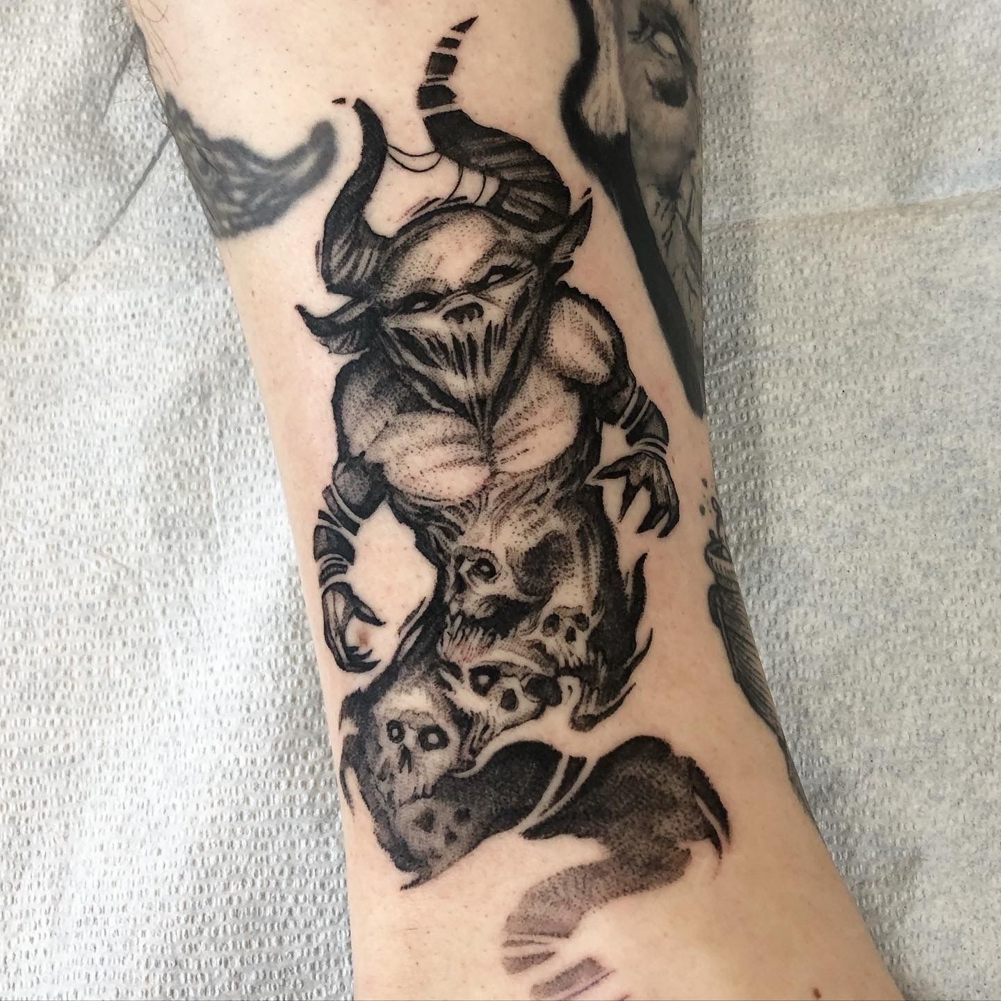 Black-and-gray tattoo of a horned creature with skull-like face on a forearm