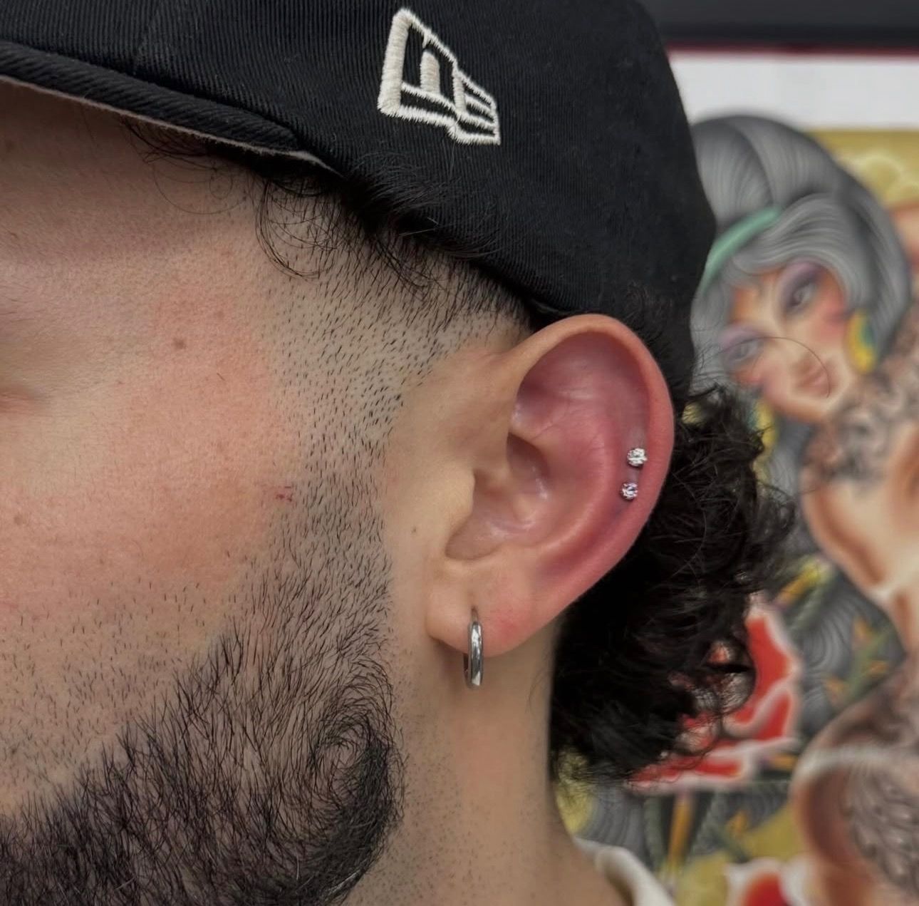 Close-up of a man’s ear with hoop and stud piercings, wearing a black cap, with a tattoo in the background