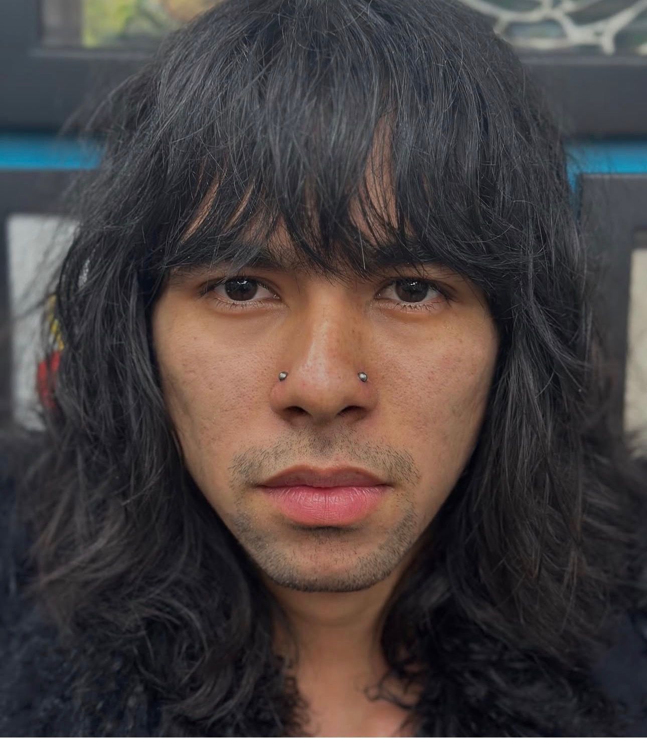 Close-up portrait of a person with long dark hair and bangs, neutral expression, nose piercings visible.