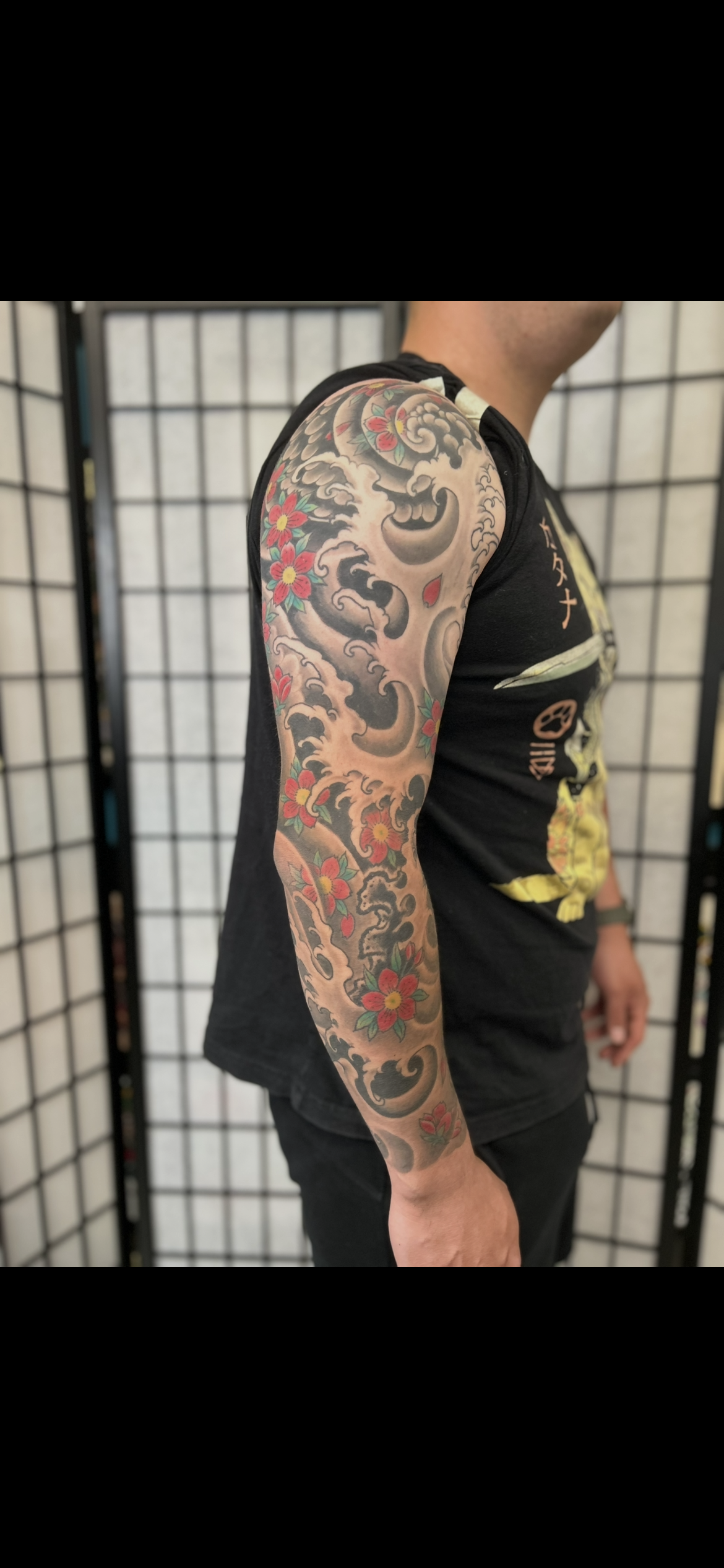 Person in black tank top showing a colorful full-sleeve tattoo on their arm against a folding screen backdrop
