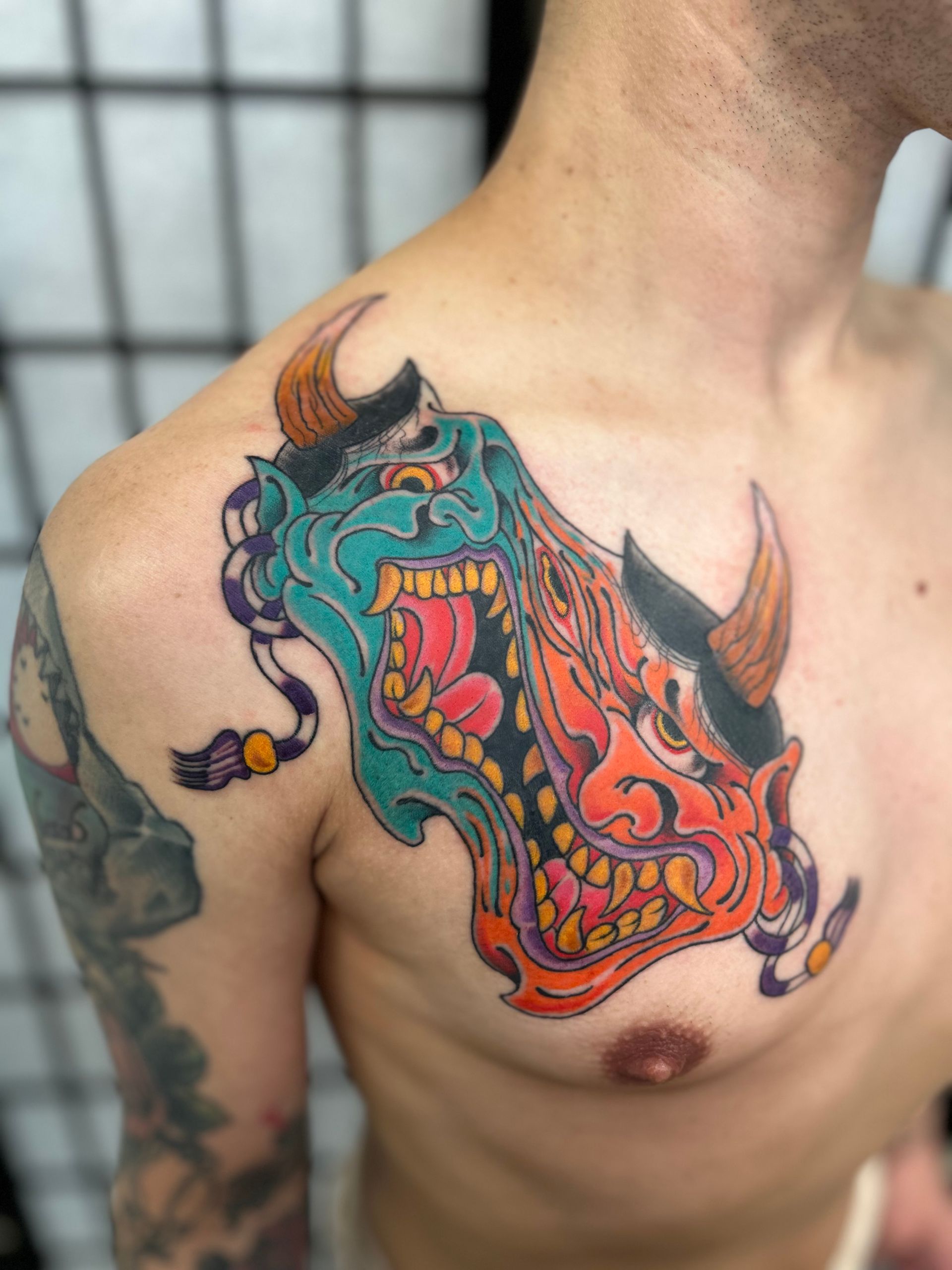Colorful horned demon tattoo on a person’s upper chest and shoulder, with teal, orange, and purple details