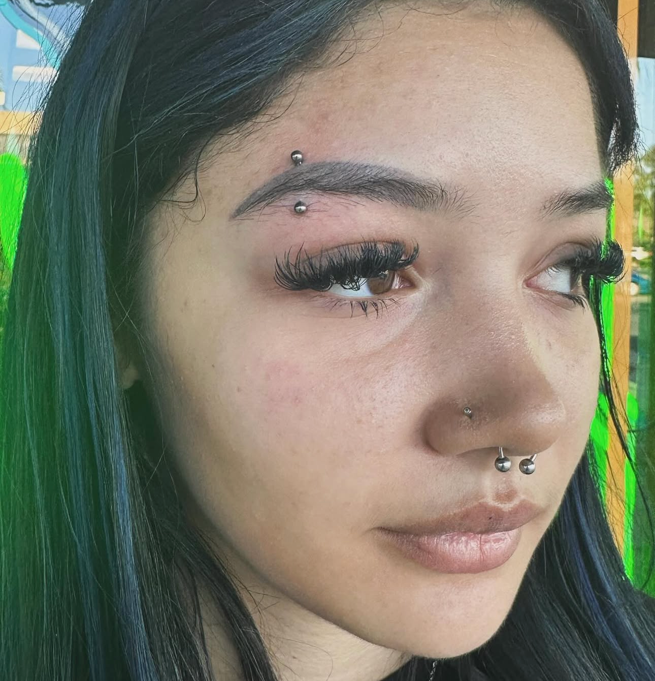 Close-up of a person with dark hair, eyebrow and nose piercings, looking to the right.
