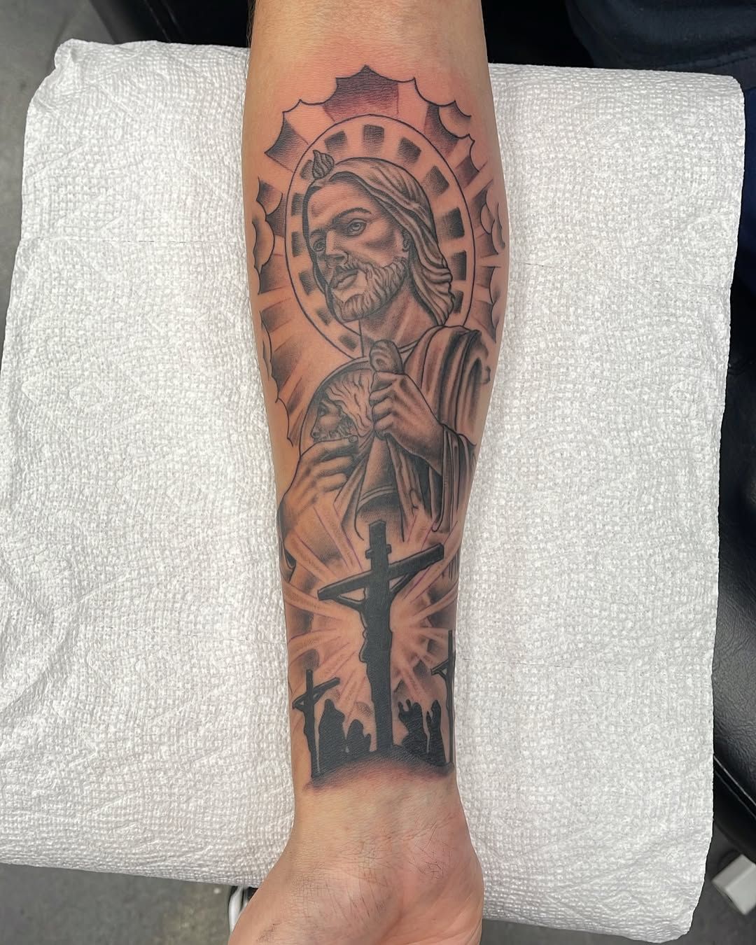 Forearm tattoo of a praying figure with a halo and cross, in dark black-and-gray ink.