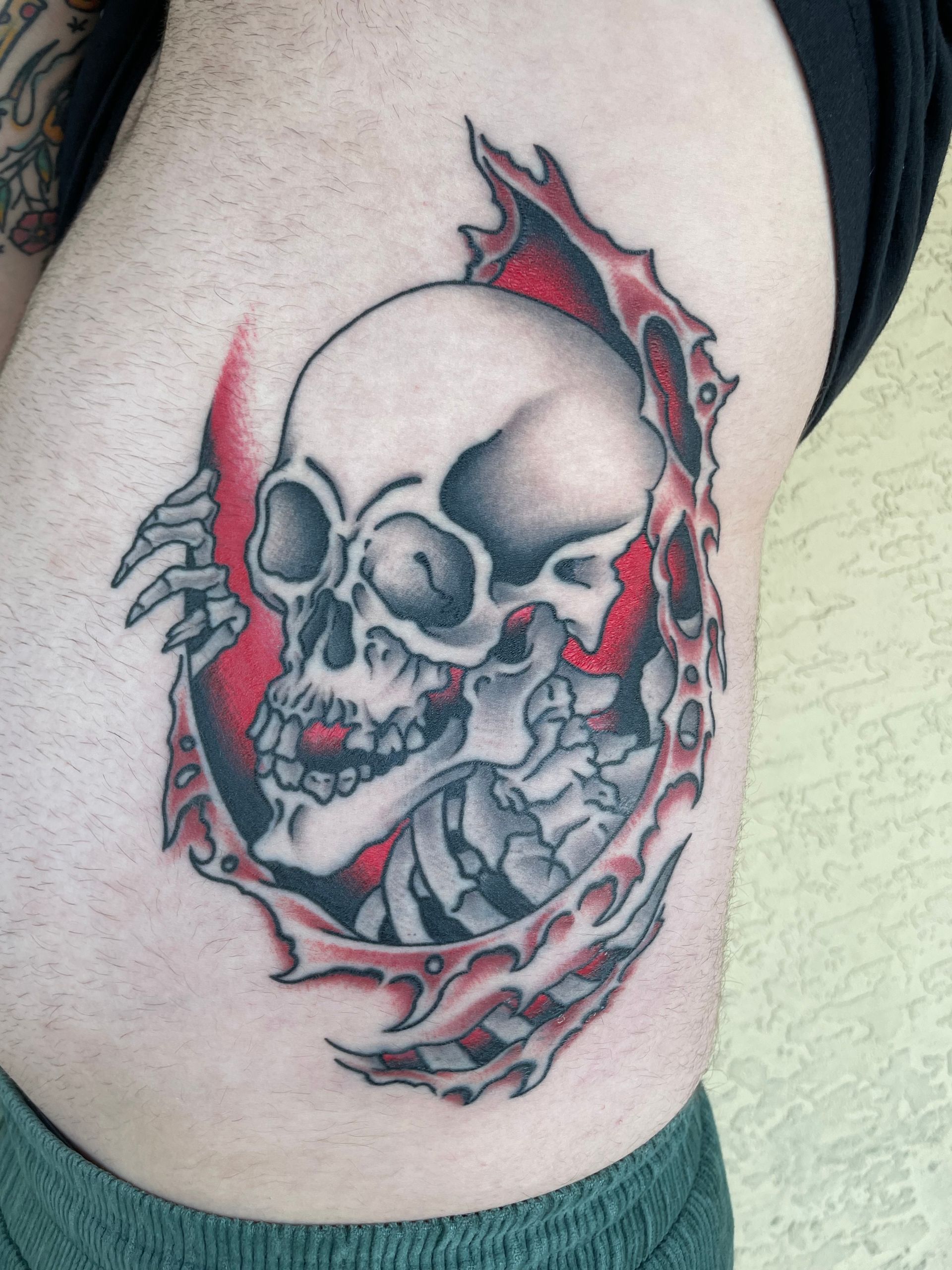 Skull tattoo with red and gray shading, framed by a torn circular design on a person’s side
