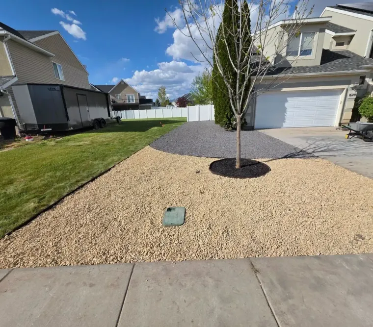 Front yard with gravel landscaping, a young tree, and a suburban house with a white garage door.