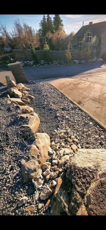 Rock border beside a gravel path and paved driveway in a landscaped yard with trees and a house in the background