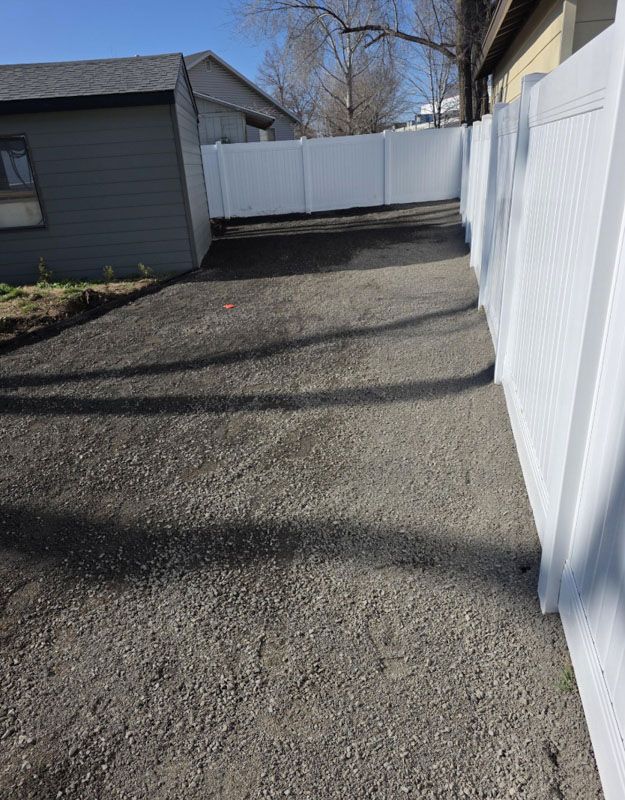 Narrow gravel driveway beside white vinyl fence and gray garage on a sunny day