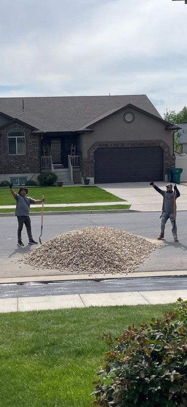 Two people stand beside a large gravel pile in the street in front of a suburban house.