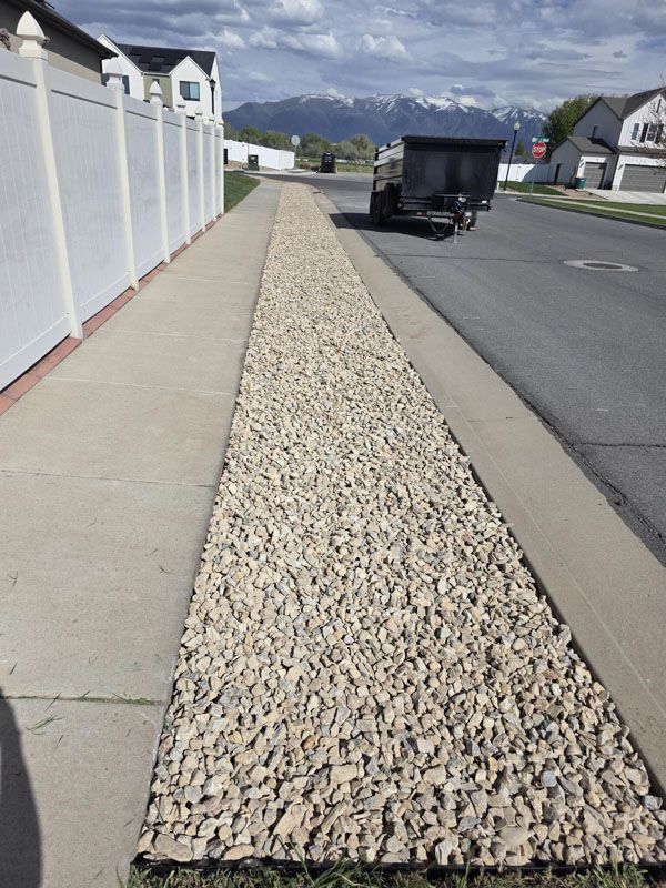 Landscaped rock strip between a sidewalk and street beside a white fence, with a dumpster ahead.