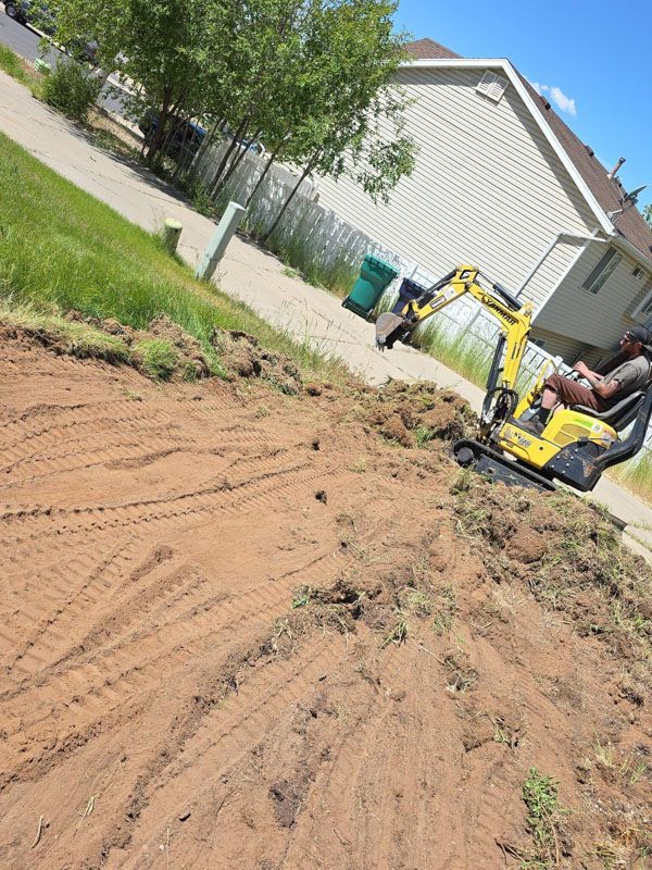 Construction worker using a yellow excavator on a dirt lot beside a house and tree on a sunny day
