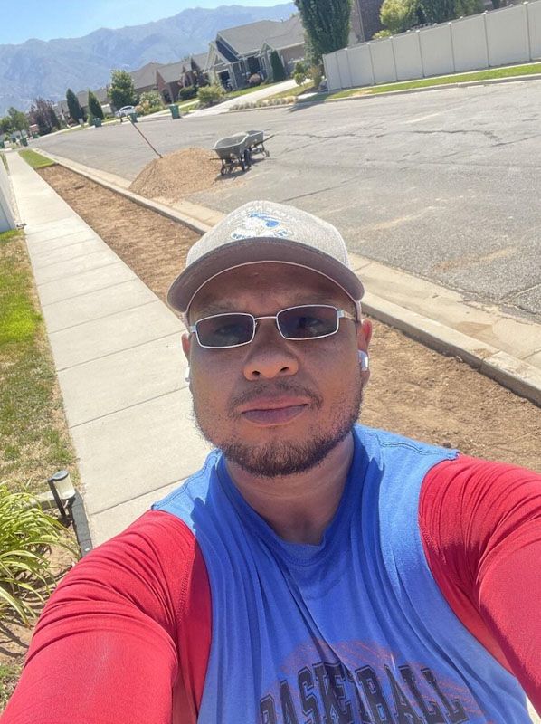 Selfie of a person in a red-and-blue shirt and cap beside a sidewalk on a sunny roadside