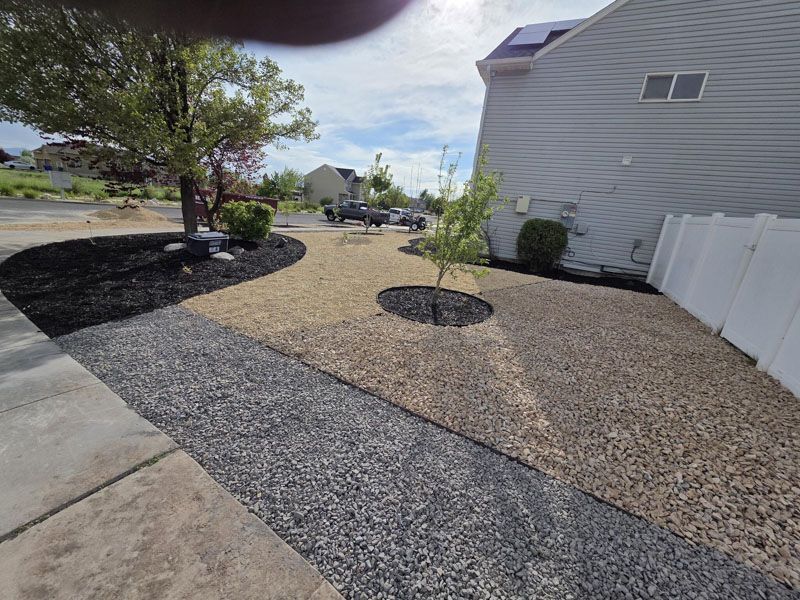Landscaped side yard with gravel beds, young trees, concrete walkway, and a white fence beside a house.