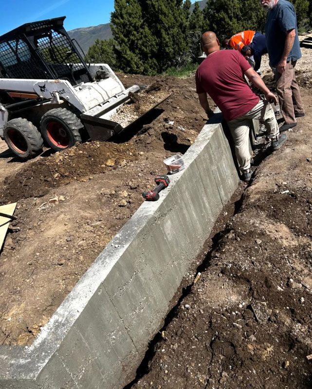 Workers beside a concrete retaining wall under construction with a skid steer and dirt excavation.