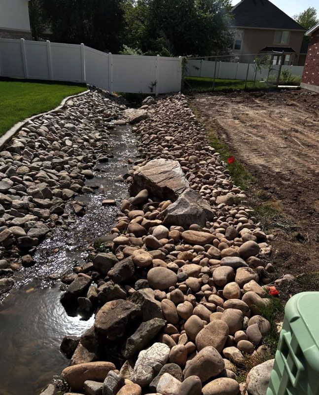 Rock-lined drainage ditch with flowing water beside a grassy yard and a dirt bank