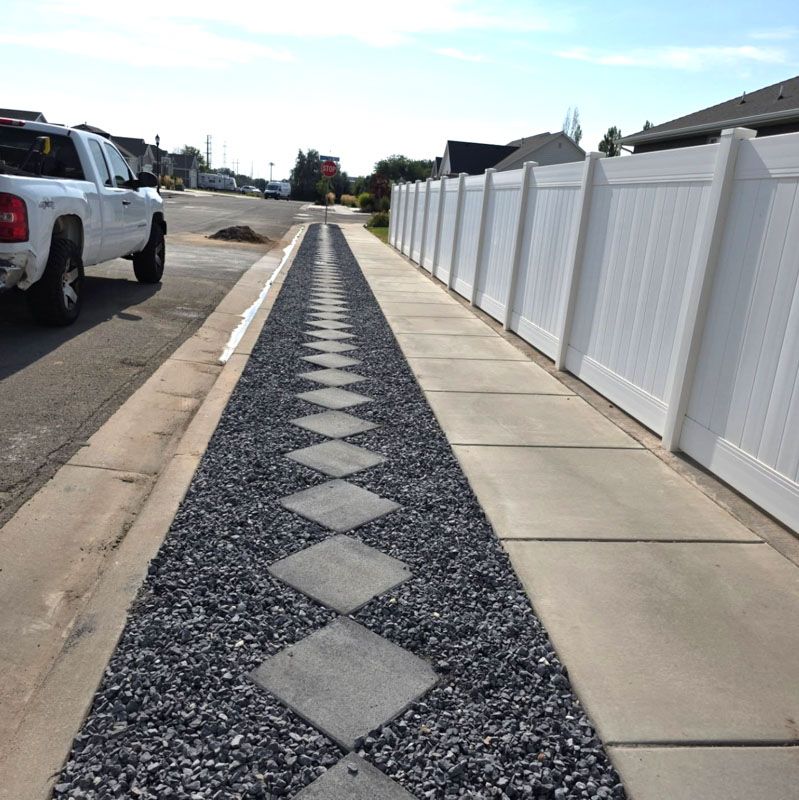 Concrete sidewalk with a gravel center strip and diamond pattern beside a white fence and parked truck.