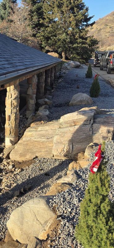 Stone-lined gravel walkway beside a rustic building, with trees, boulders, and a red fire extinguisher.