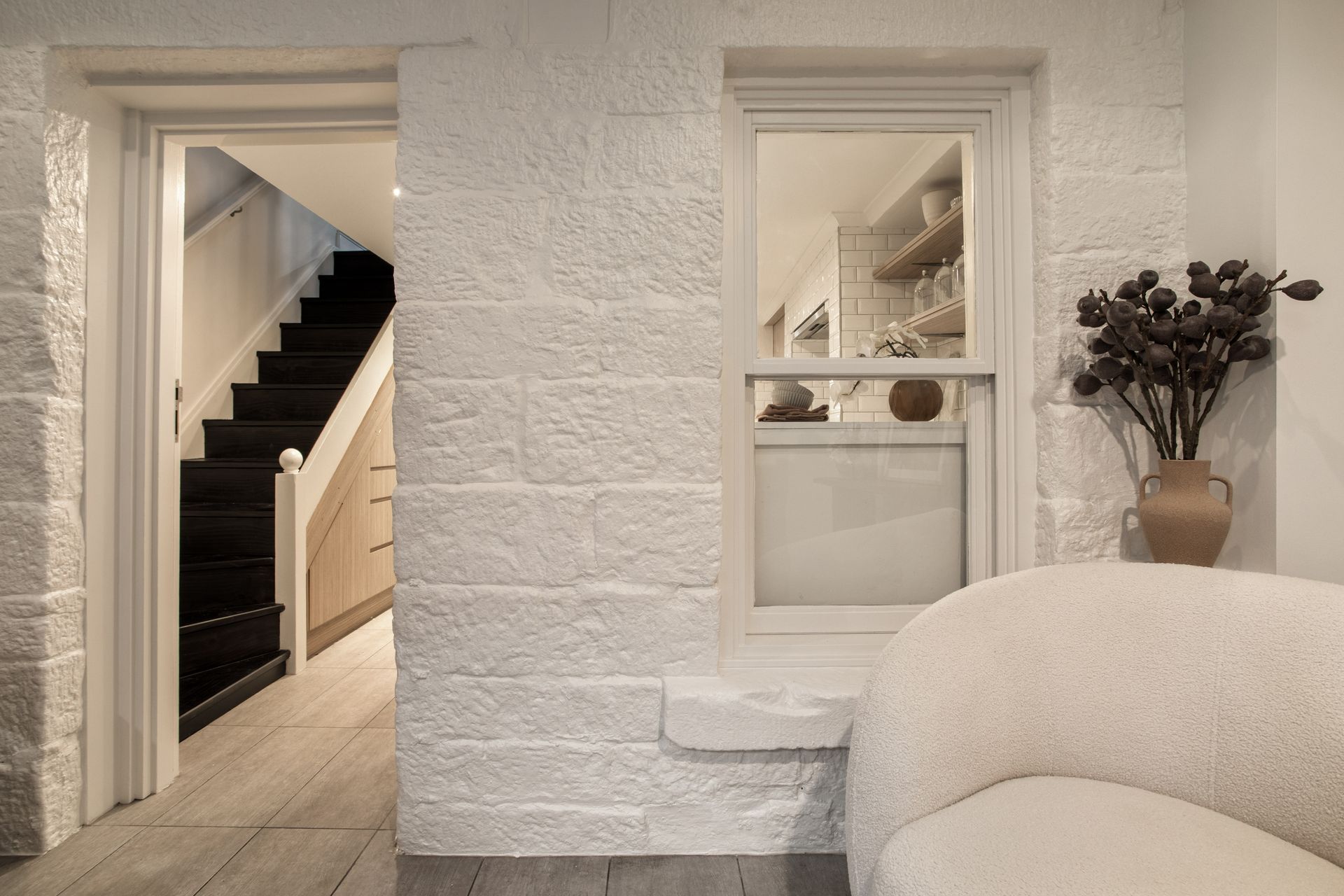 Bright hallway with open doorway, staircase, white textured walls, and a vase of dried flowers by a sofa