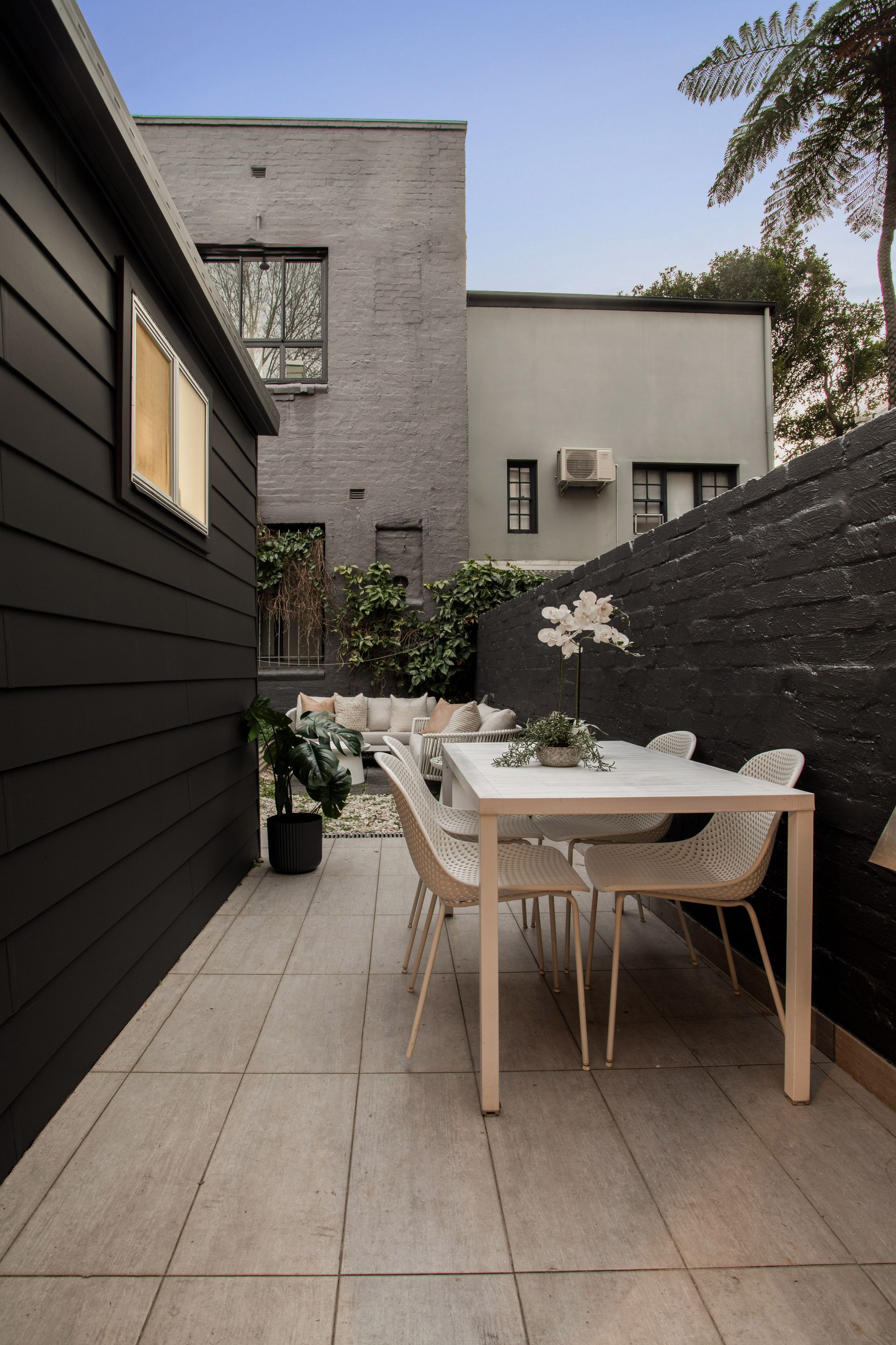 Narrow outdoor patio with a white table and chairs, potted plants, and brick walls beside a modern home.