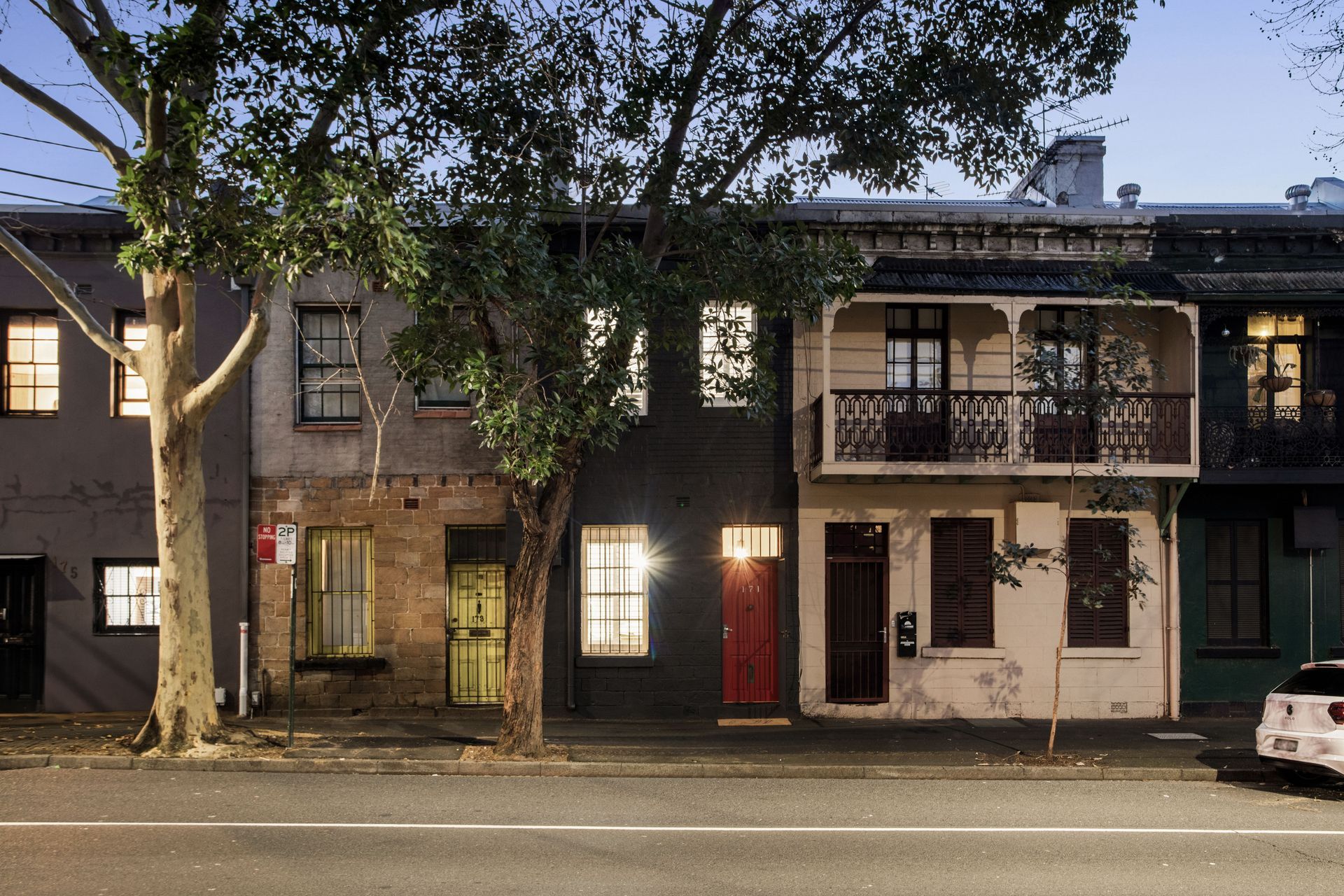 Row of lit townhouses at dusk with trees along the street and a red front door