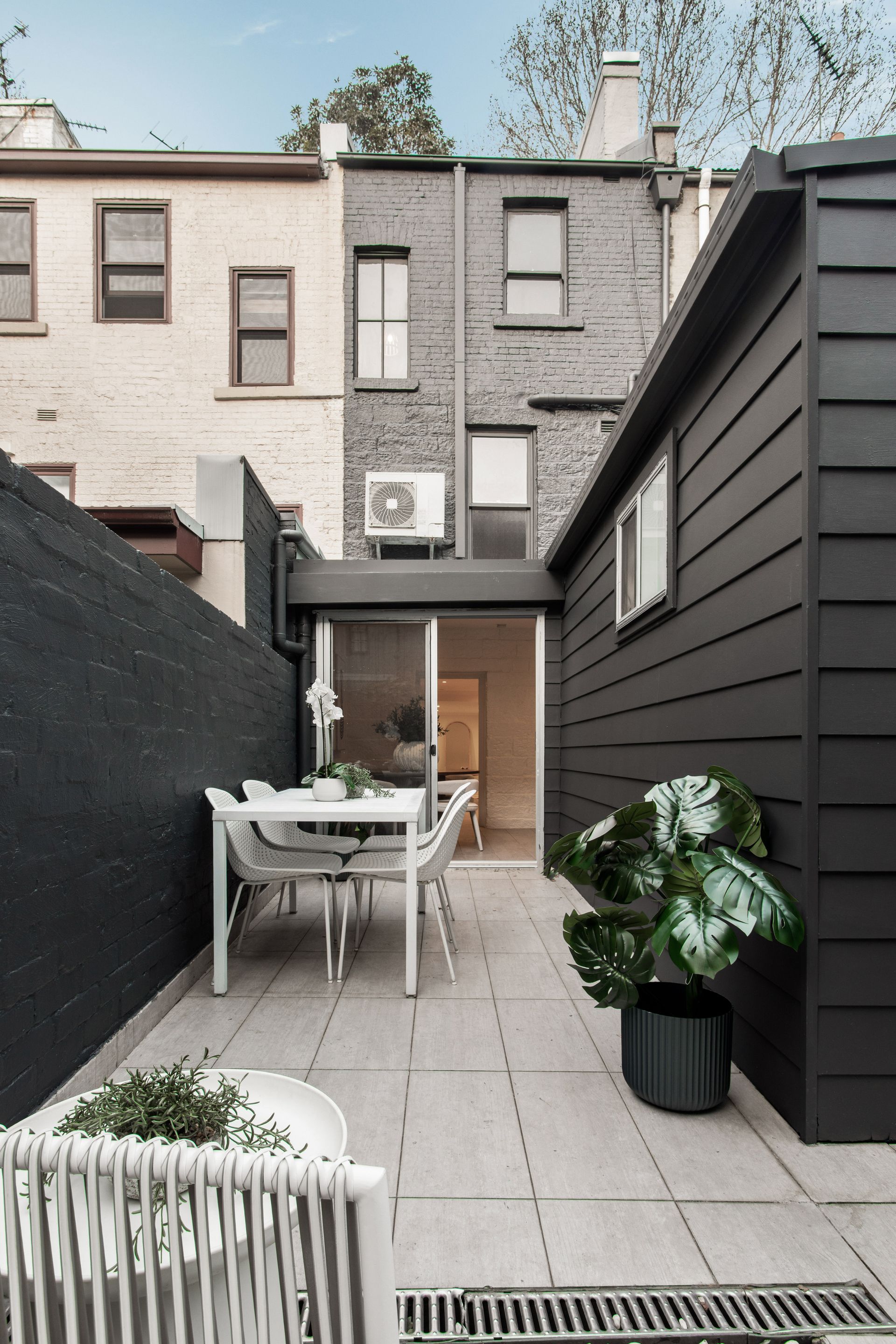 Narrow paved courtyard with white table and chairs, potted plant, and dark wood siding beside brick buildings.