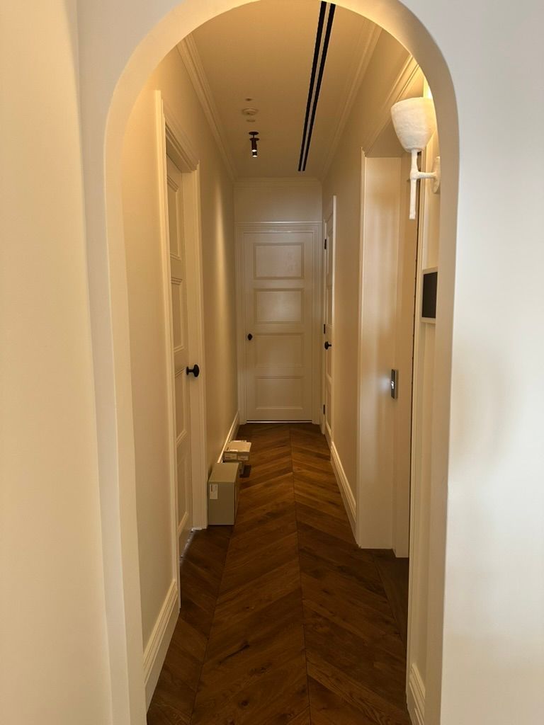 Narrow arched hallway with beige walls, herringbone wood floor, and a white door at the end