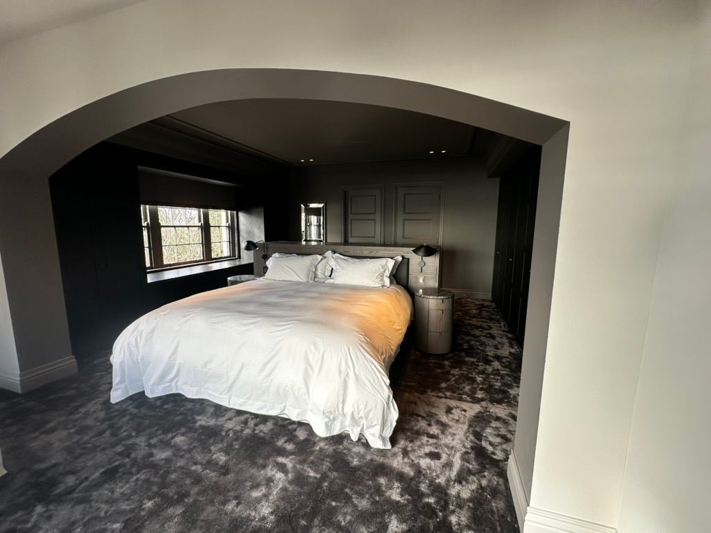 Bedroom with a white bed and gray carpet, viewed through a wide archway into a dark, cozy room