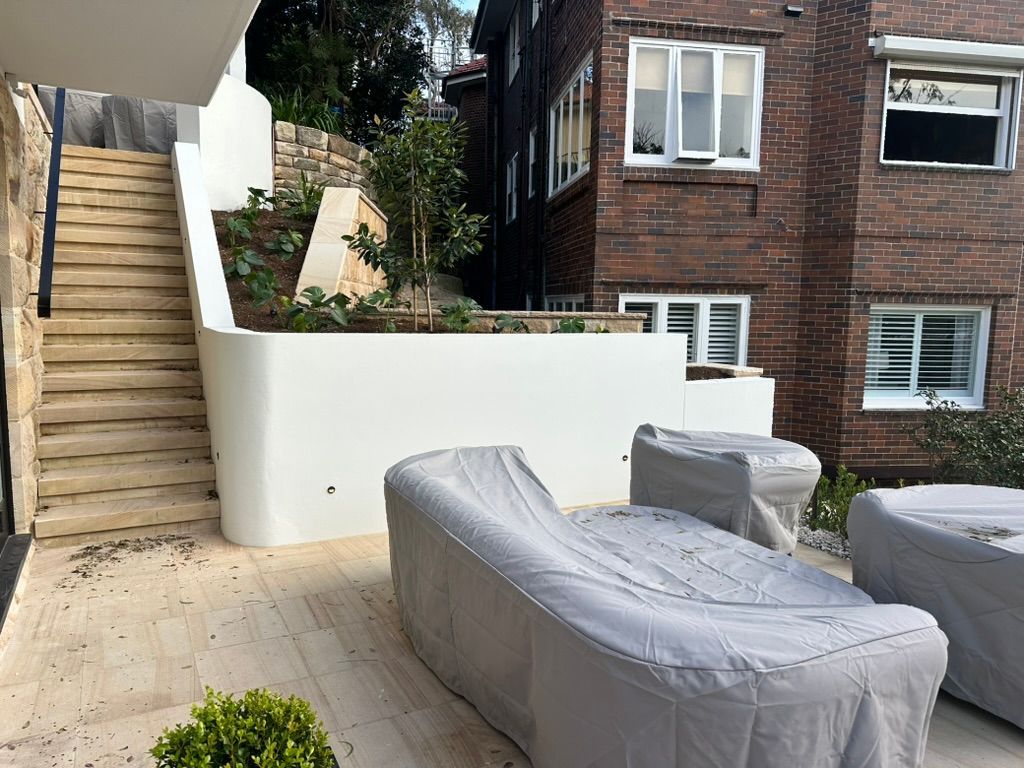 Outdoor patio with covered lounge chairs, stairs, and a brick building beside a white retaining wall