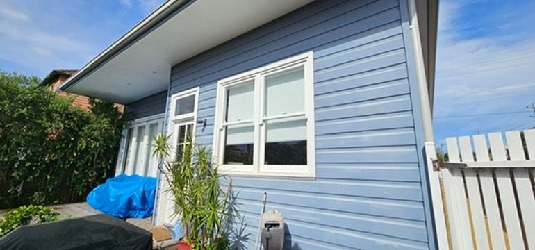 Exterior of a blue house with white-trimmed windows, a white fence, and a covered side walkway
