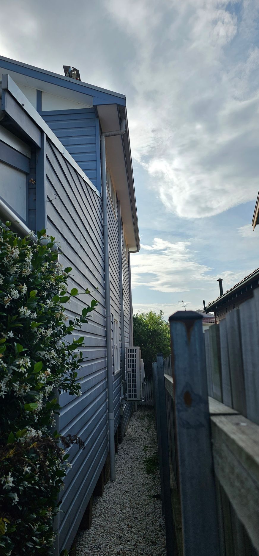 Narrow side yard between blue house and fence under cloudy sky, with shrubs and a small outdoor unit.