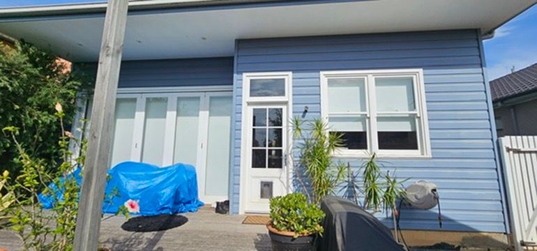 Blue house front with white door and windows, covered chair on porch, and potted plants by the entrance