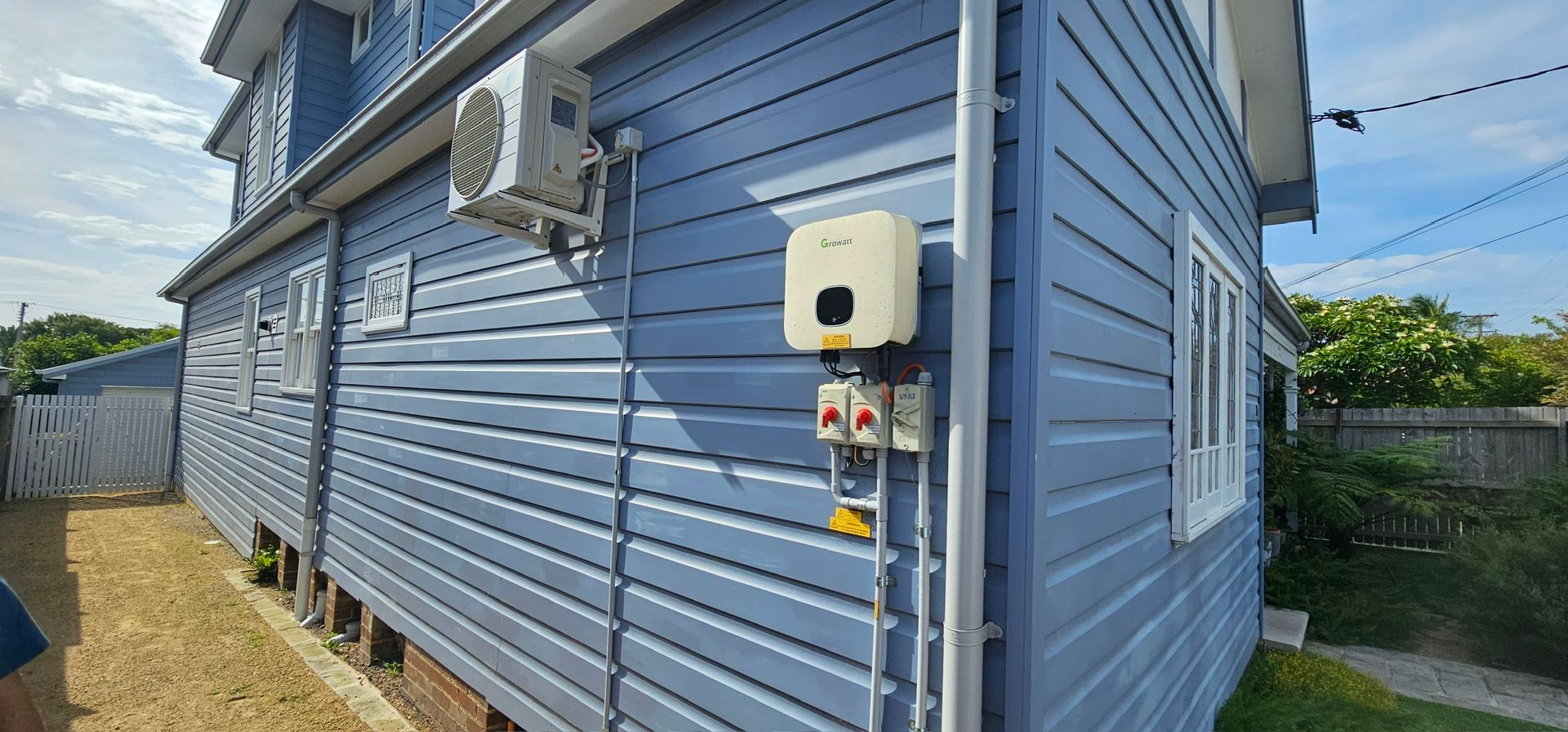 Blue house exterior with side-mounted utility boxes, air conditioner, and fenced yard under a sunny sky