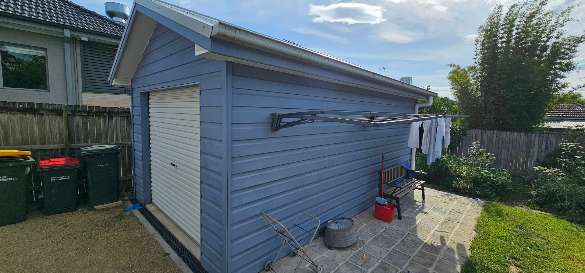 Blue garage with a white door in a backyard, with patio items and a bicycle rack nearby.