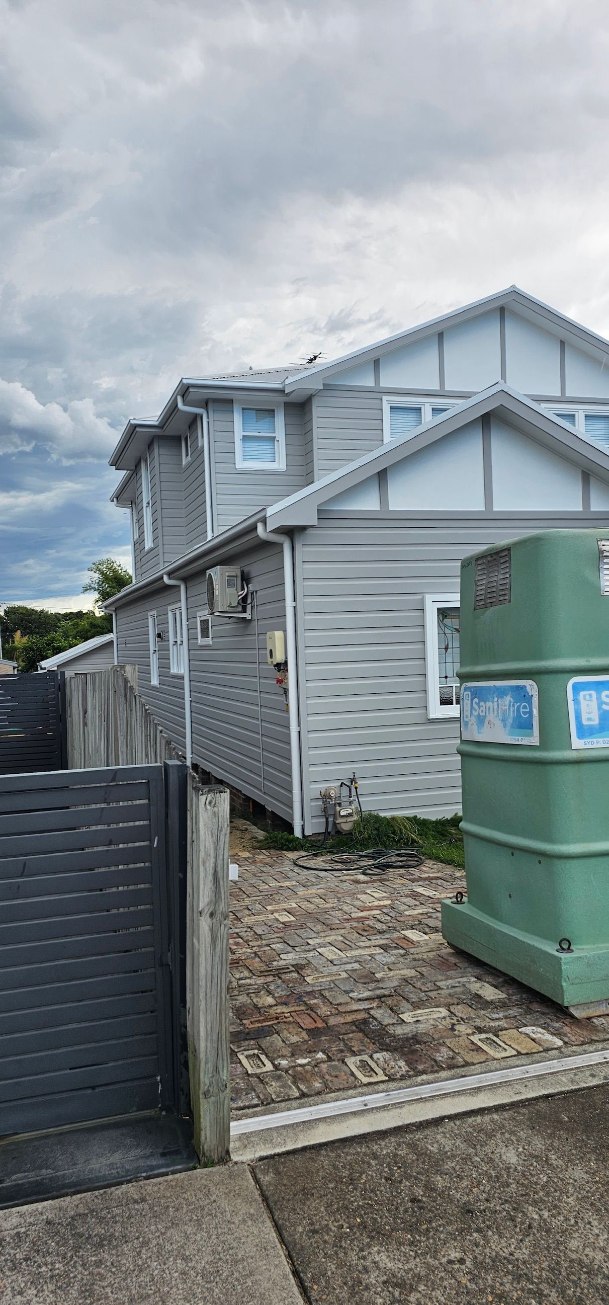 Gray two-story house with a covered side porch, black fence, and green storage tanks under cloudy skies