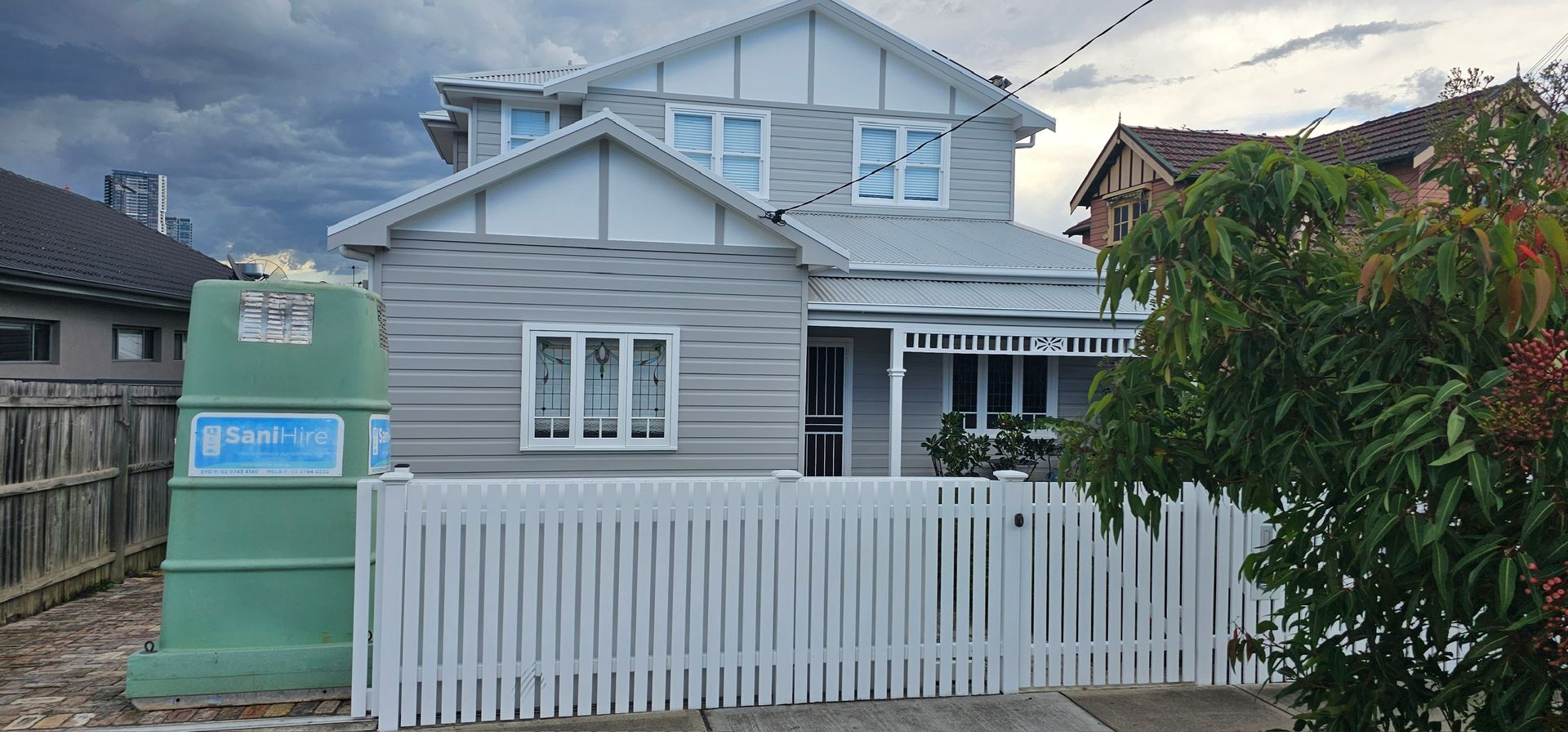 Gray house with white fence and porch, beside a green dumpster on a residential street