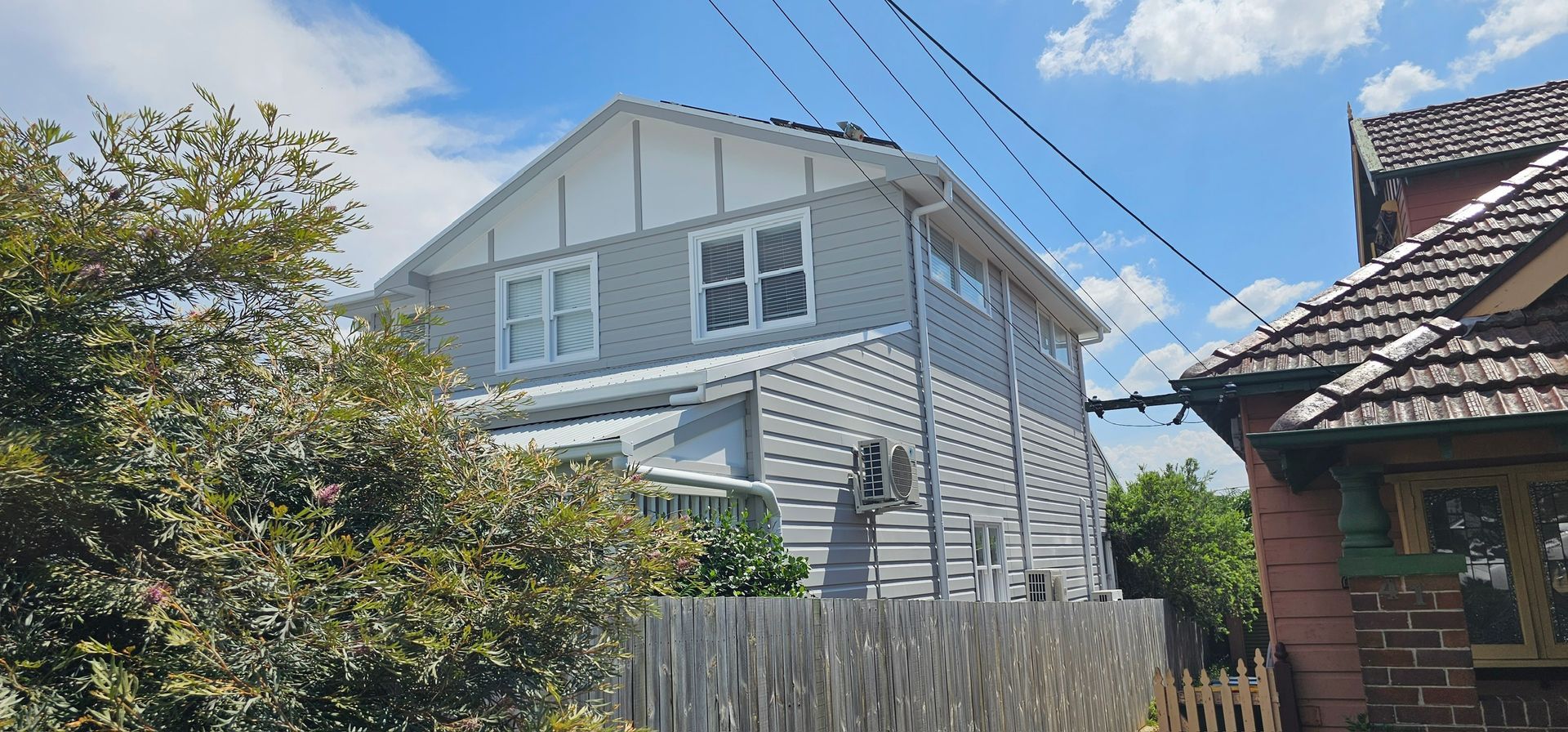 White two-story house with gray siding, blue sky, trees, and a wooden fence in the foreground