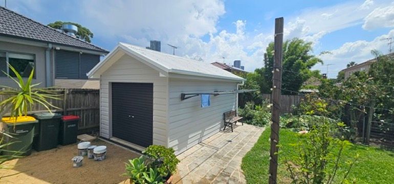 Backyard view of a small white garage with a black door beside a garden and palm trees