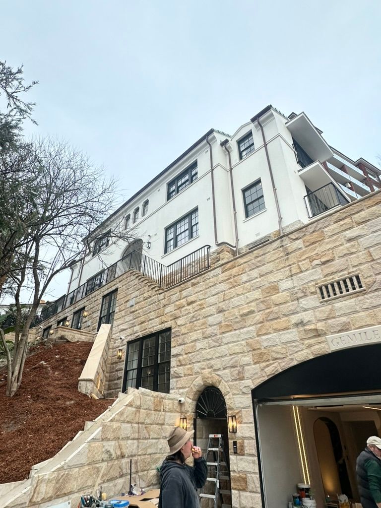 Stone building facade on a steep hillside, with a white upper section and a person standing near the entrance.
