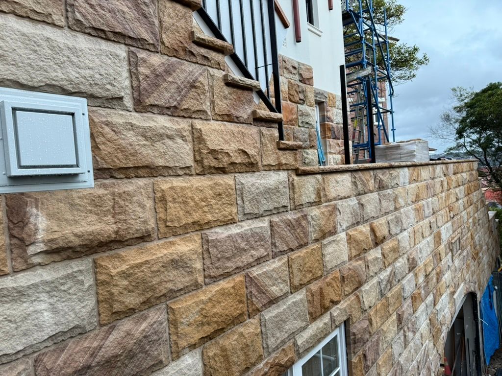 Stone retaining wall beside a house, with stairs, windows, and a cloudy sky above