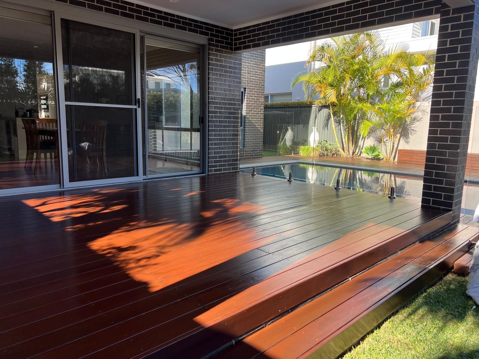 Covered wooden deck with glass railing and brick posts beside a grassy yard