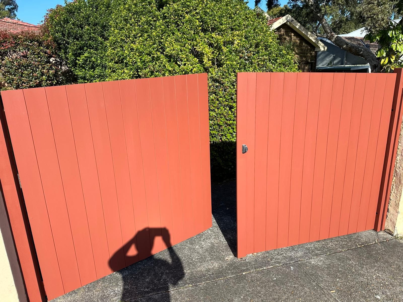 Open red wooden gates leading to a sunny backyard with bushes and a shadowed driveway.