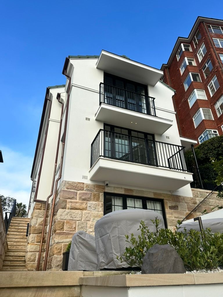 Modern white house with balconies and stone foundation on a hillside, beside red brick buildings and blue sky