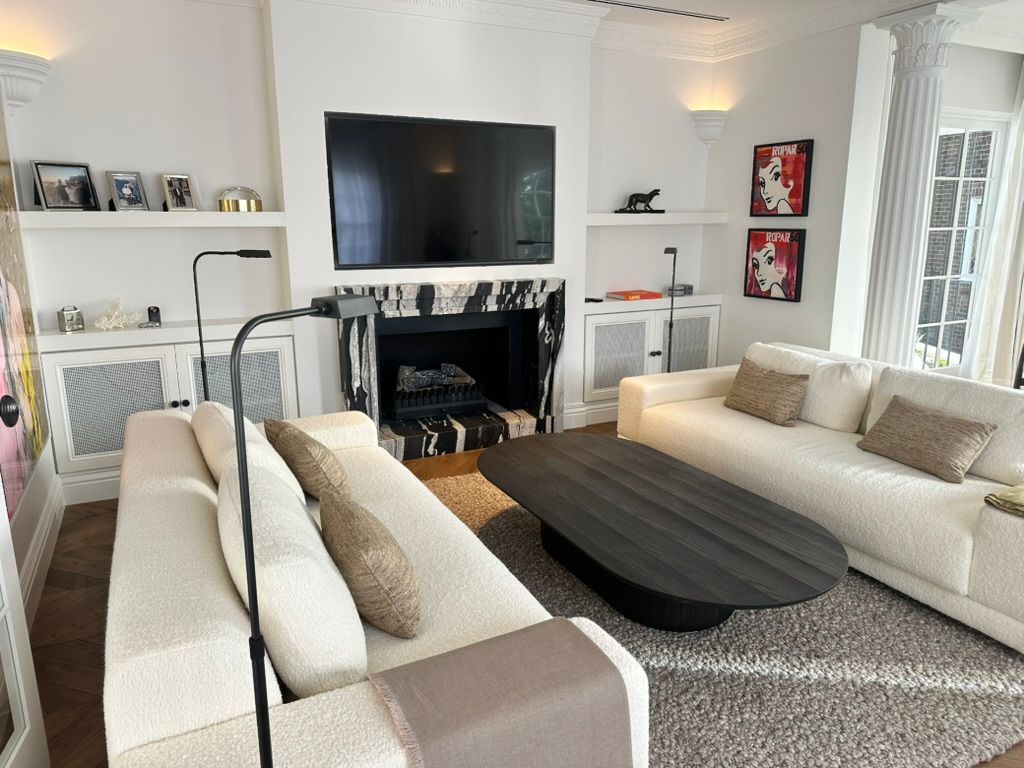 Bright living room with two white sofas, black coffee table, TV above fireplace, and large windows