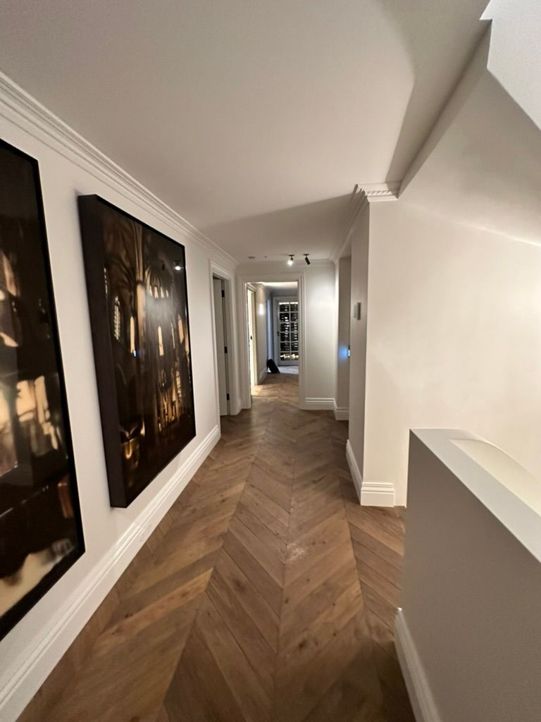Long hallway with herringbone wood floors, white walls, and framed artwork on the left.