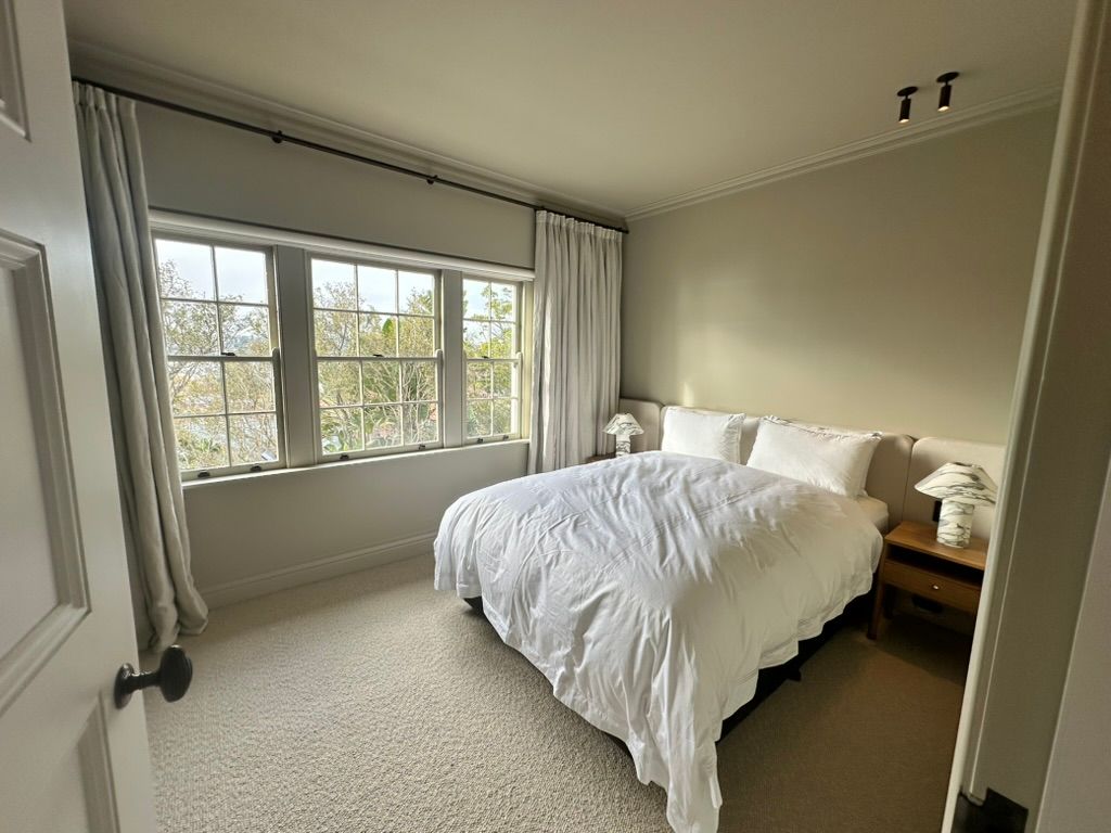 Bright bedroom with a large window, white bedding, beige walls, and a wooden nightstand