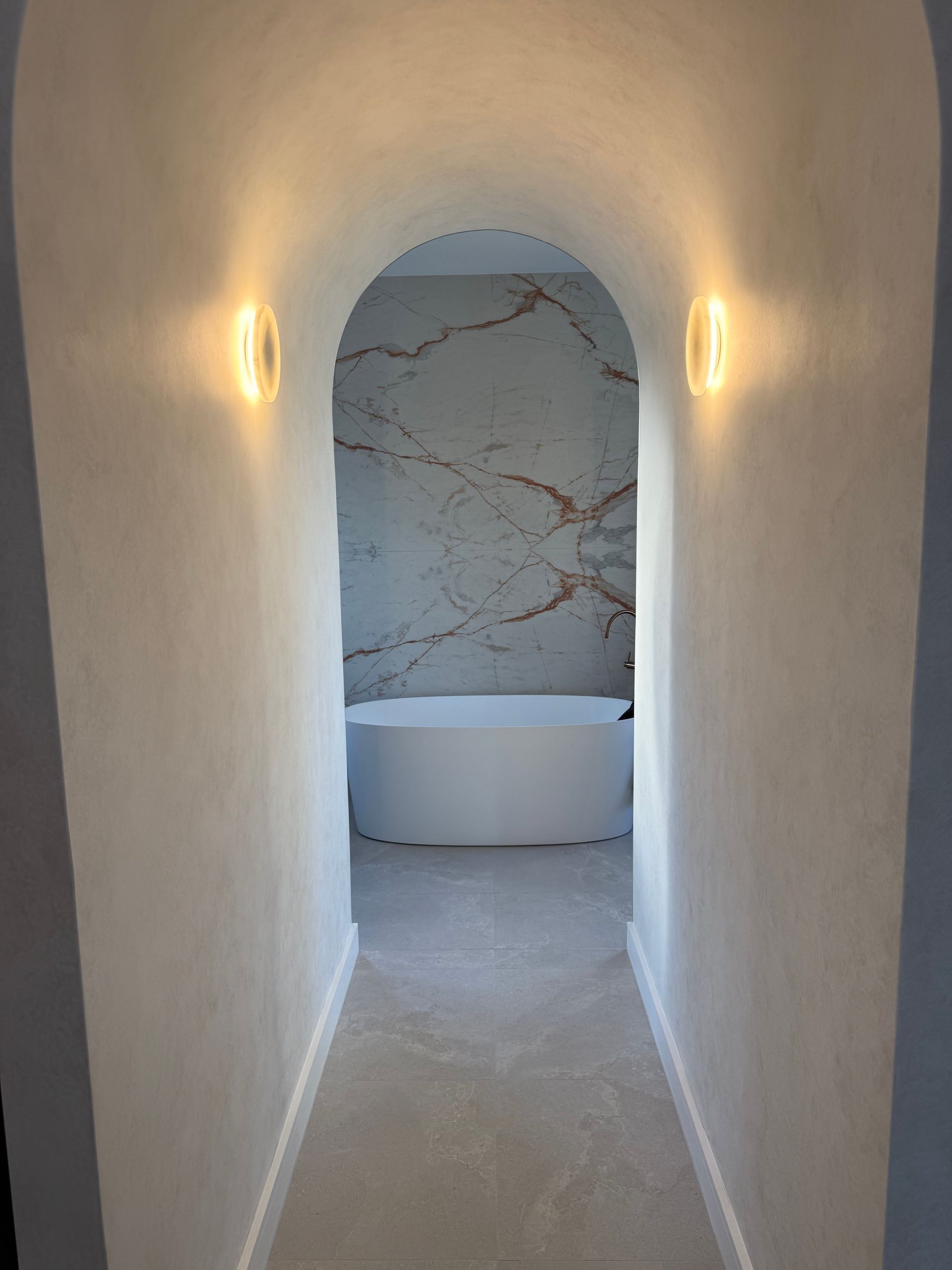 Narrow white hallway with wall sconces leading to a marble bathroom and freestanding tub