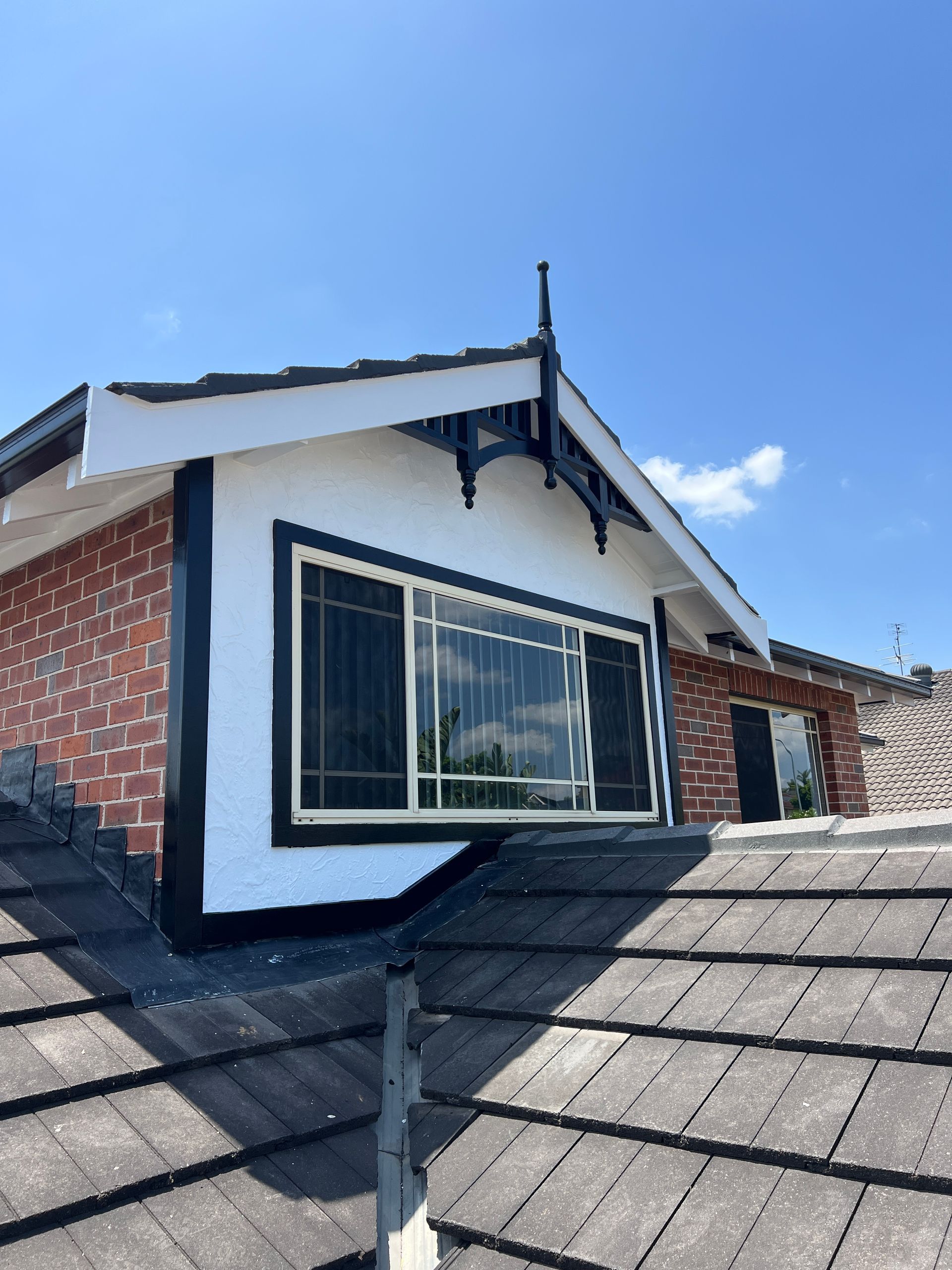 Roof dormer with white siding, black trim, and a large window on a shingled rooftop under blue sky