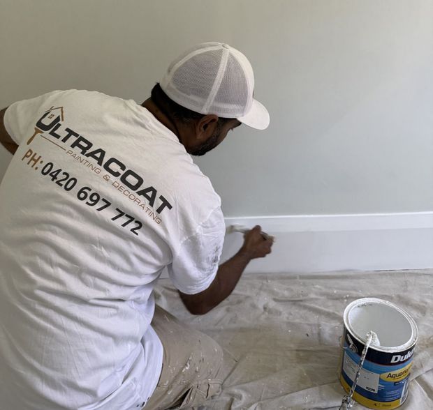 Painter in white shirt and cap painting a white wall beside a bucket on a covered floor.