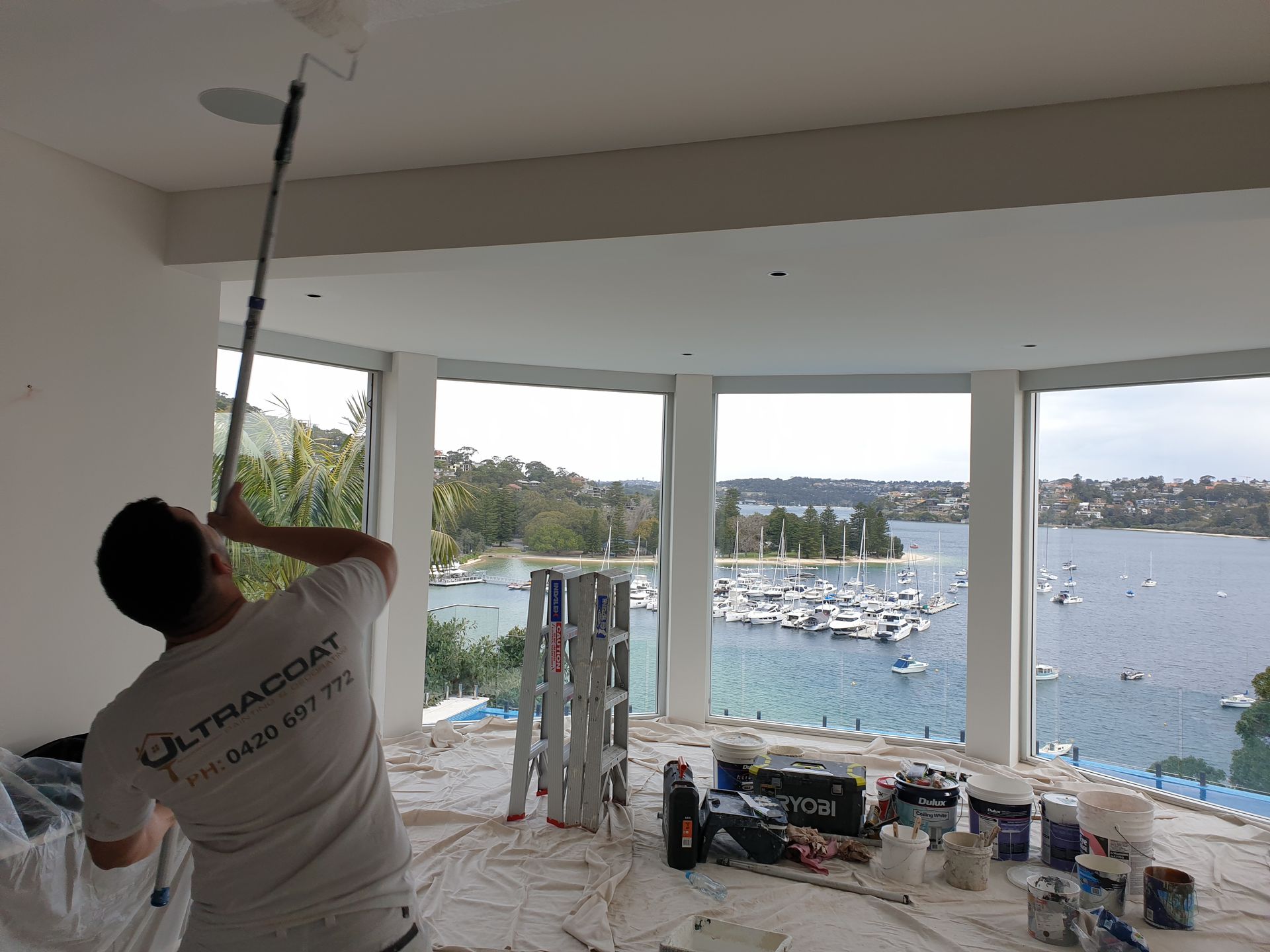 Worker painting a ceiling inside a room with large windows overlooking a marina. Worker painting a ceiling inside a room with large windows overlooking a marina.