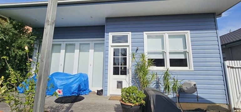 Blue house front porch with white-trimmed windows, a door, potted plants, and a tarp-covered object.